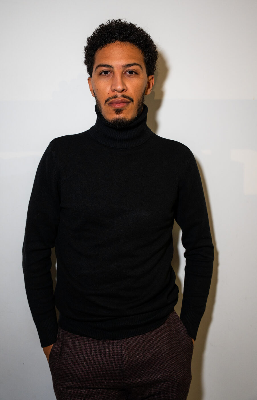 Hamza Bouzhar standing against a white wall in a black turtleneck, hands in his pockets.