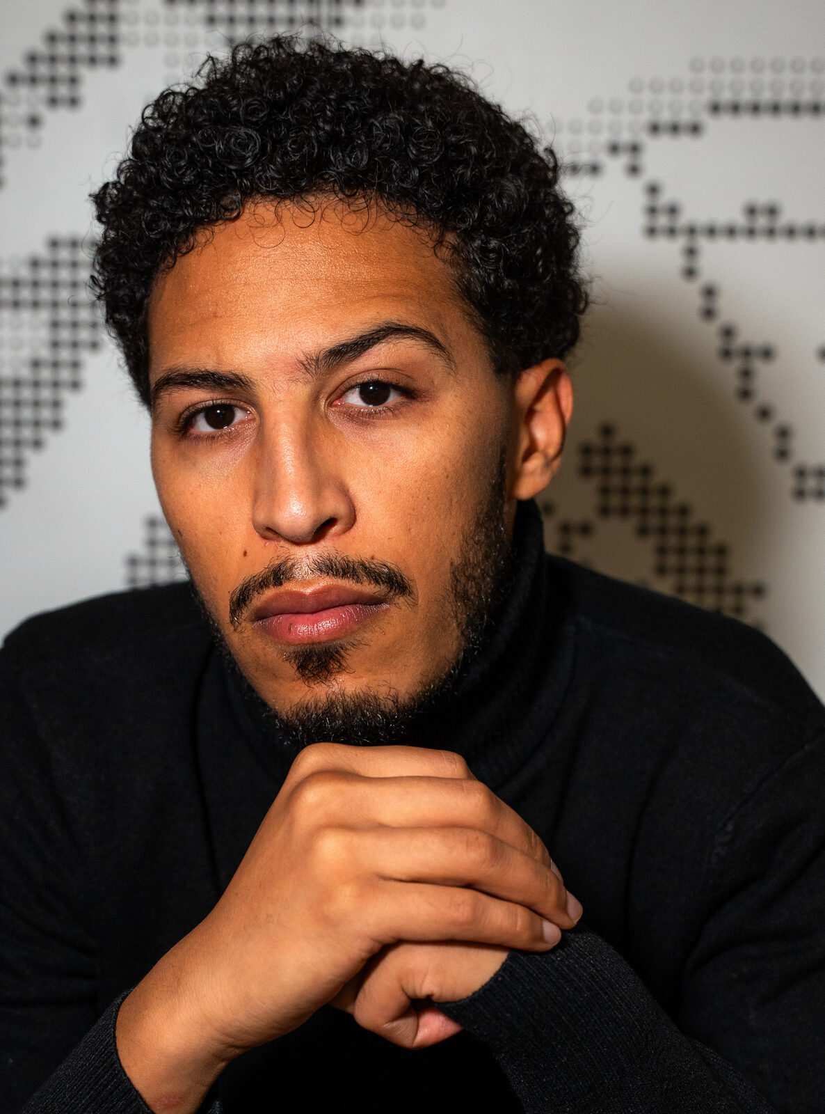 Hamza Bouzhar in a black turtleneck, hands clasped under his chin, in front of a dotted map wall.